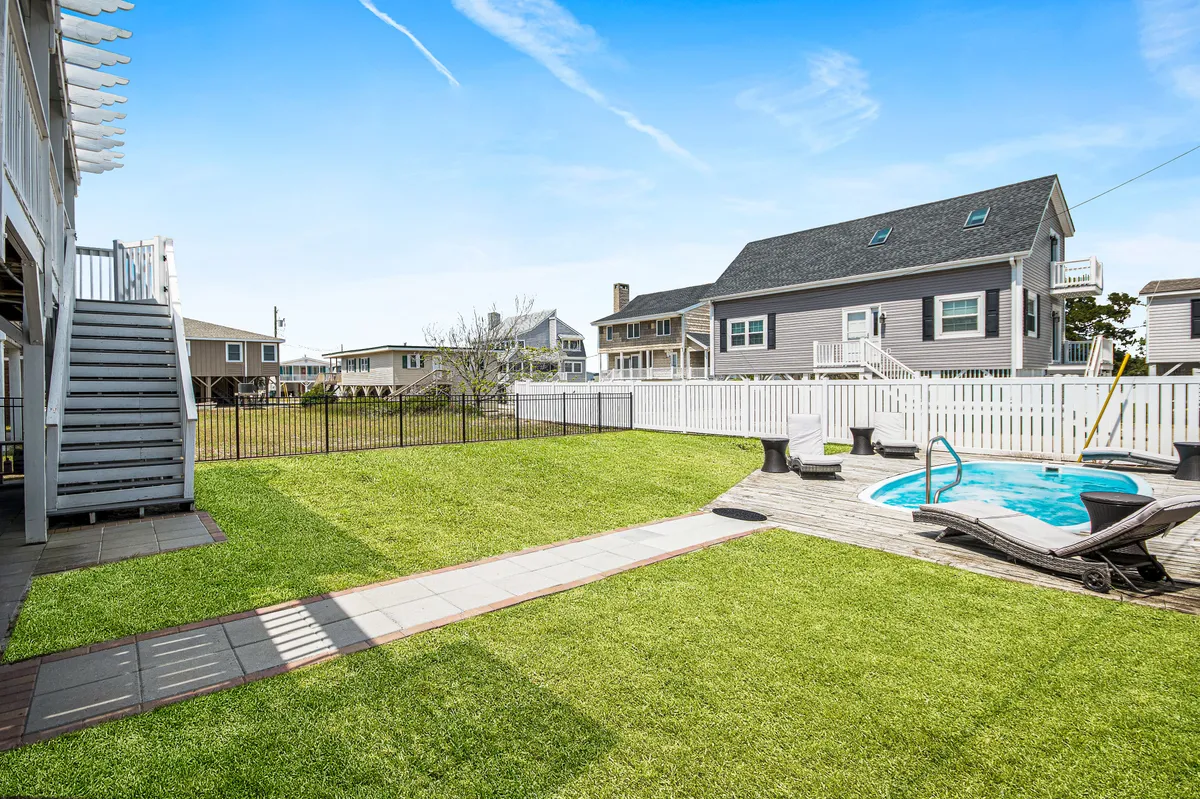 A bright and inviting backyard features a sparkling swimming pool surrounded by a green lawn and a spacious patio area. Patio furniture and lounge chairs are arranged for sunbathing and relaxing, while a sturdy fence encloses the yard, making the area both private and secure. The scene is cheerful and ideal for family gatherings or poolside relaxation.