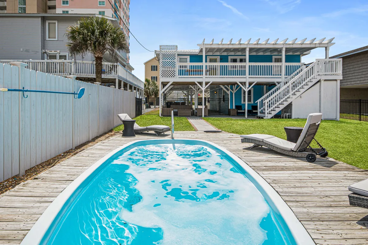 A serene backyard scene showcases a well-sized swimming pool surrounded by several lounge chairs, a wooden deck, palm trees, and a view of the home's structure. The area is ideal for both sunbathing and social gatherings, with clear skies and lush landscaping enhancing the tropical ambiance.