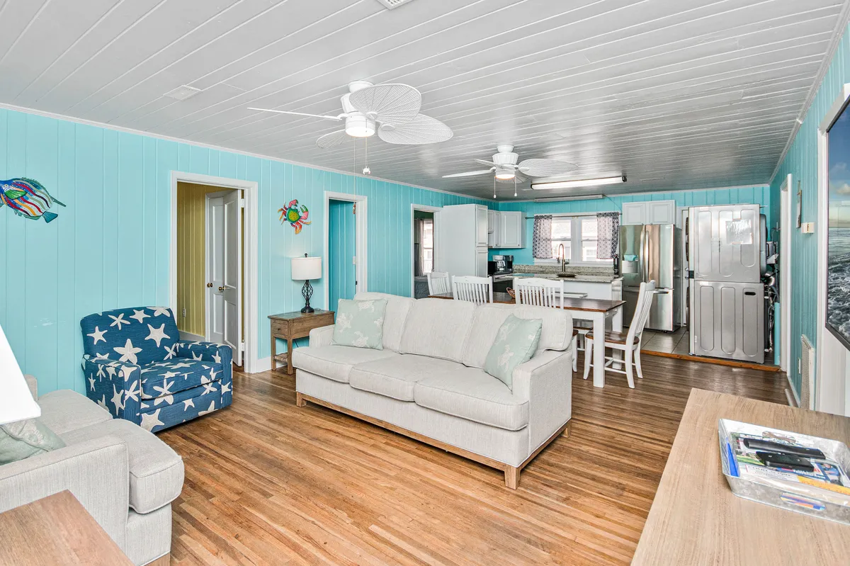 A bright and inviting open-concept living and dining area with light blue walls, a comfortable sofa, a star-patterned accent chair, and a dining table set for a group meal. Natural light fills the space, highlighting the casual coastal décor and plush seating.