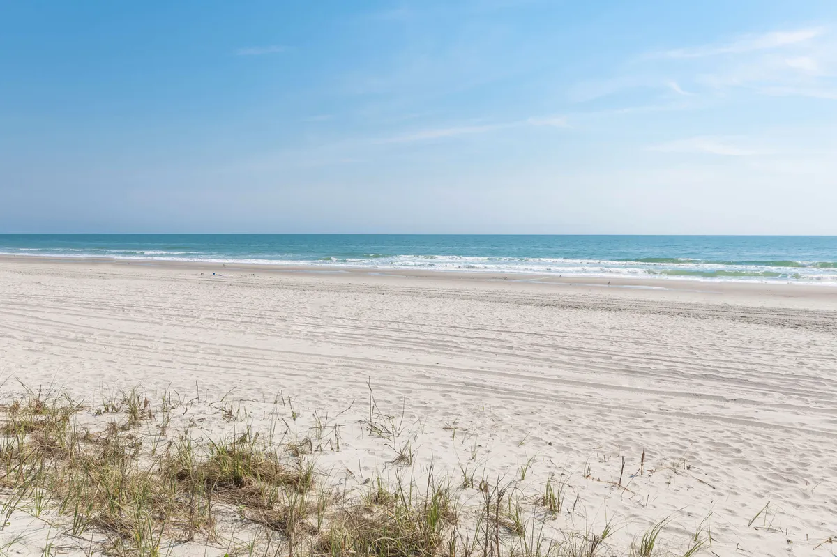 A scenic view of Cherry Grove Beach with rolling sand dunes, gentle ocean waves, and a clear blue sky. The scene is tranquil, with space for sunbathing, beach walks, and play. The untouched sand and open water evoke the classic coastal experience.