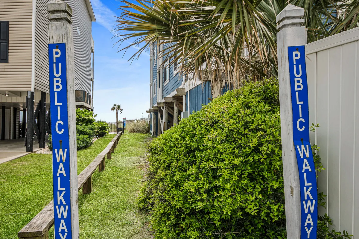 A public walkway sign is set amidst lush greenery, leading directly to the sandy pathway that provides beach access. The scene suggests a relaxing and inviting approach to the coastline, highlighting the ease of reaching the beach from the property.
