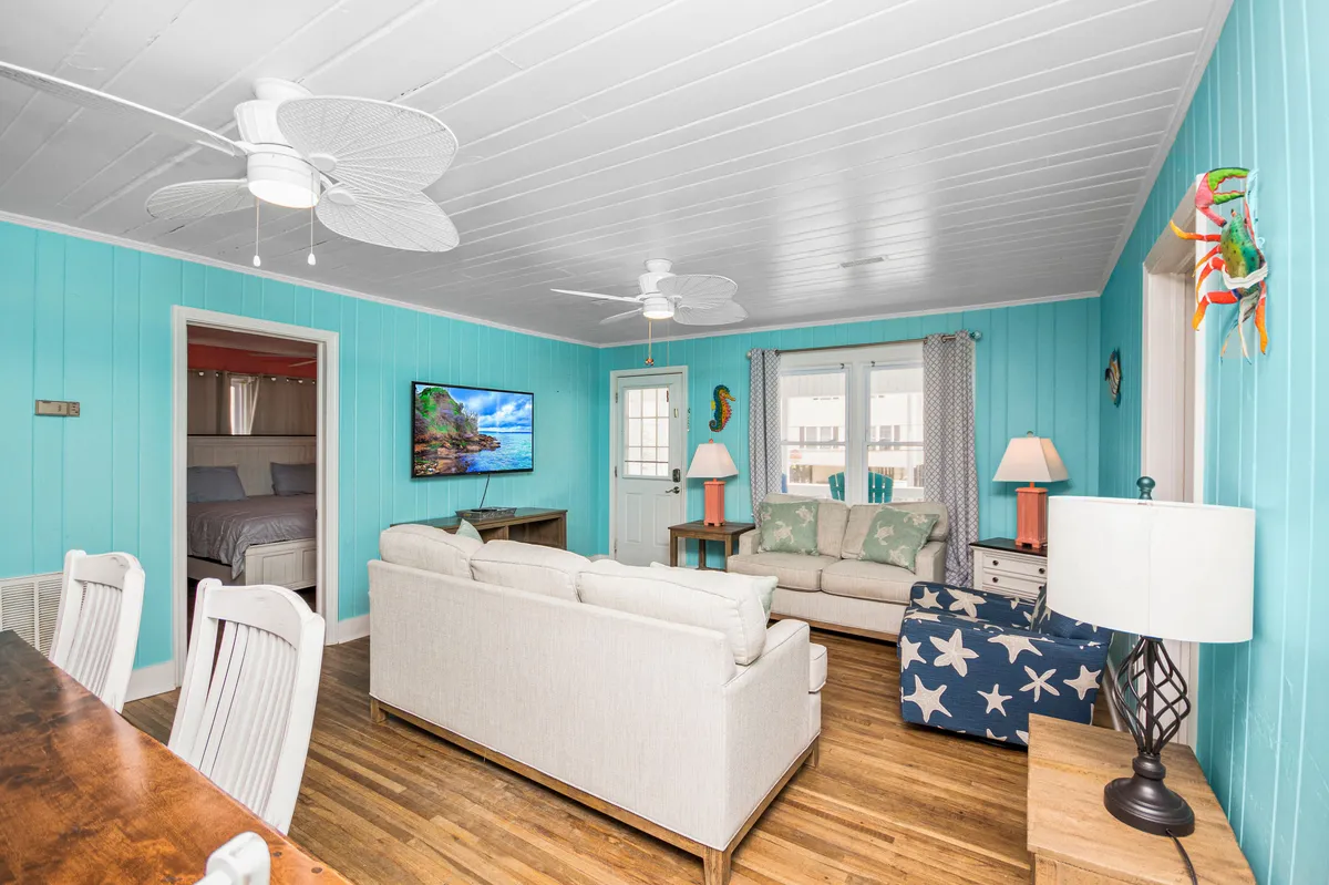A cheerful living and dining space featuring light blue walls, a comfortable sectional sofa, a TV with a scenic view onscreen, a decorative lamp, and a dining table set for a group. The room is filled with natural light, creating a welcoming backdrop for family activities.