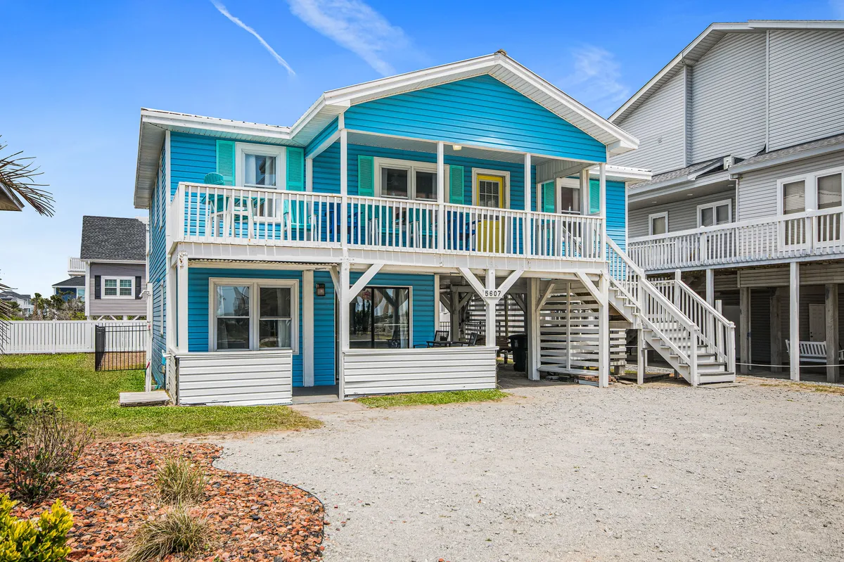 The bright blue exterior of The Salty Mermaid features white railings and a welcoming front porch. Family members are visible relaxing, with the beach nearby. The setting is bathed in warm sunset light, suggesting a peaceful, inviting retreat after a day by the sea.