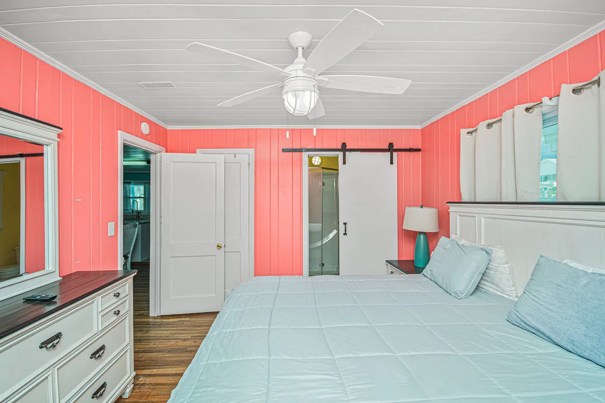 A serene bedroom with coral-colored walls, a large bed with light blue bedding, a mirror on the dresser, and a modern ceiling fan. Sunlight streams in through the windows, creating a peaceful and refreshing atmosphere. The room exemplifies coastal-inspired décor and offers a sense of space and tranquility.