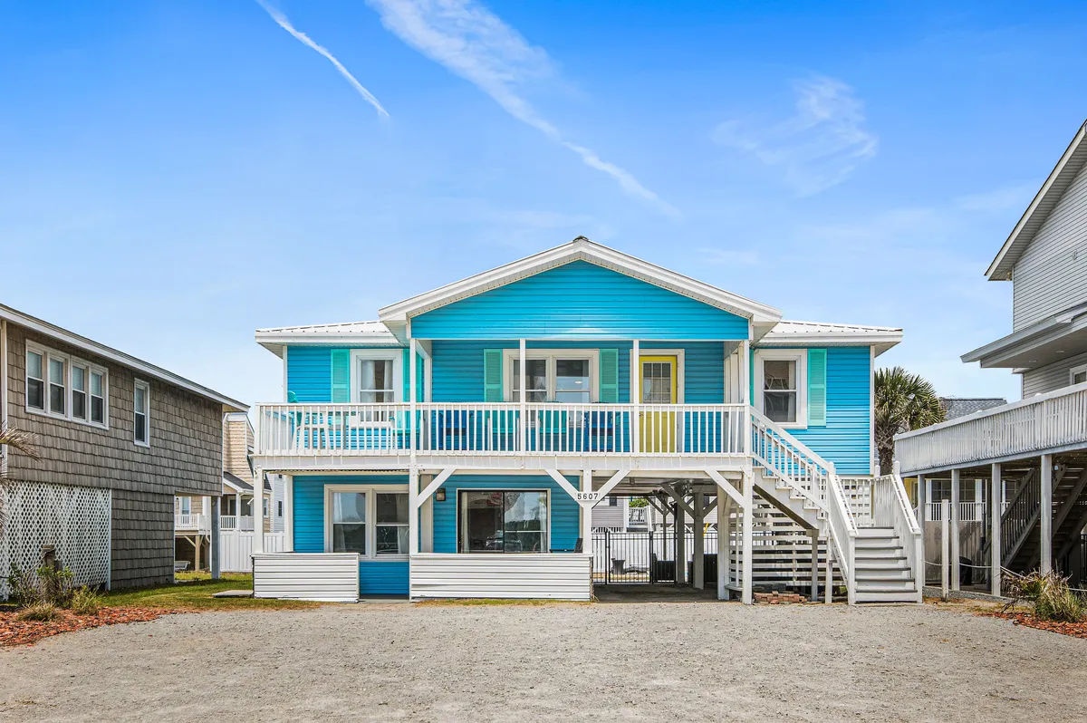 The image showcases the cheerful exterior of 'The Salty Mermaid,' a vibrant single-family house with a bright blue facade, welcoming yellow door, and white railings. The spacious porch is visible, ideal for relaxing or enjoying sunset views, and the well-kept yard suggests spaces for gatherings and play, all set just steps from the sandy beach.